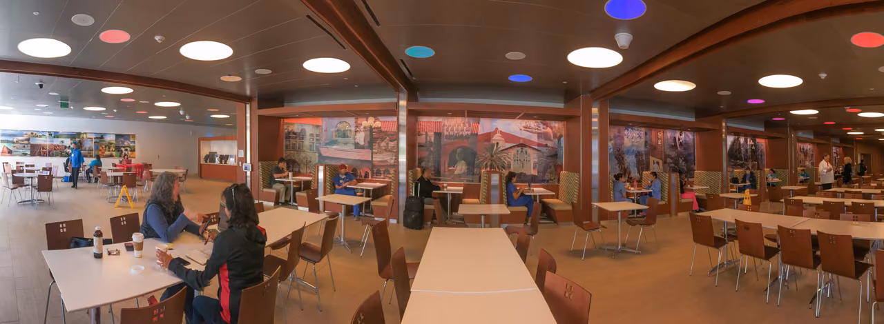 Panoramic view of a hospital cafeteria with photographic murals on the walls and diners at tables